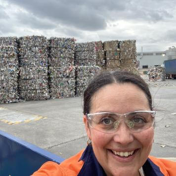 A woman wearing safety glasses and an orange high vis safety top smiles at camera, behind her is a concrete area with bales of recycled paper