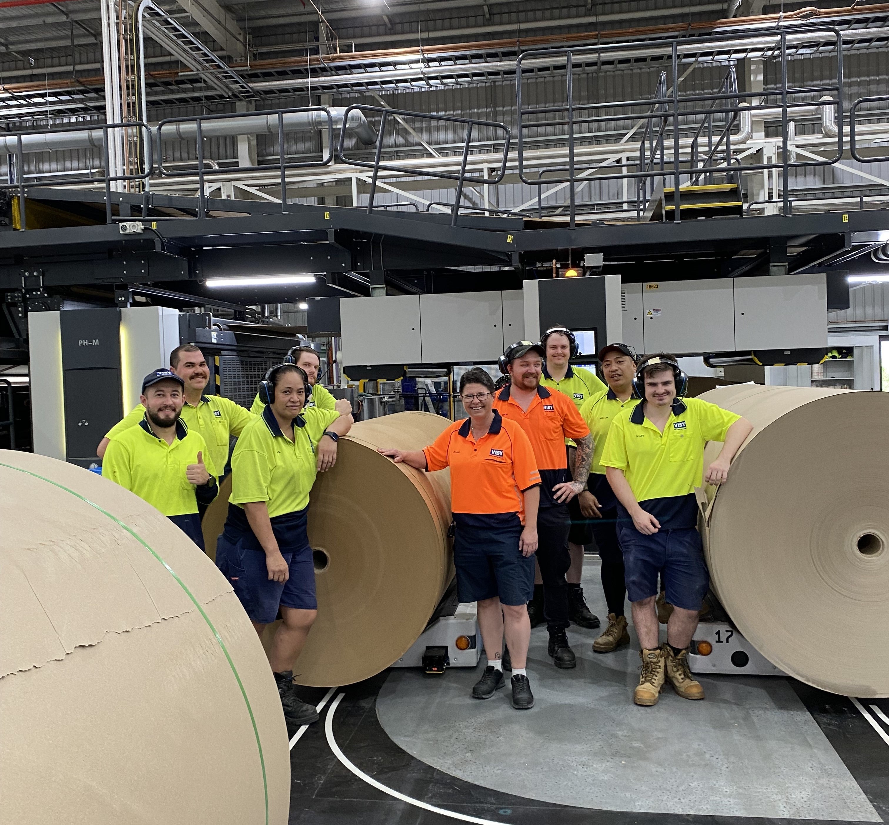 Nine people in high visibility safety vests, standing between paper reels are smiling at the camera
