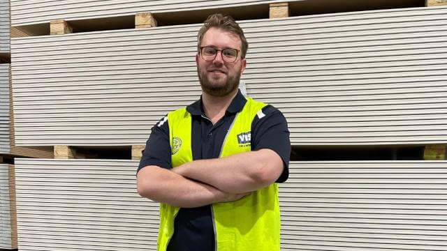 A man wearing glasses, black shirt and trousers and yellow high vis safety vest is smiling at the camera with his arms crossed. Behind him are stacks of flat pieces of board in a light tan colour. 