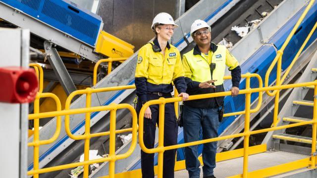 Two people smiling in high visibility safety clothes and hard hats are standing on a walk way in an industrial looking building