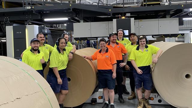 Nine people in high visibility safety vests, standing between paper reels are smiling at the camera